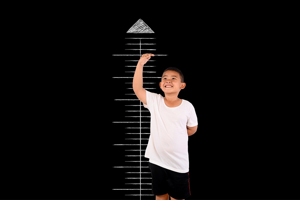 Smiling young boy in white t-shirt measuring his height against a chalk-drawn scale on a black background.