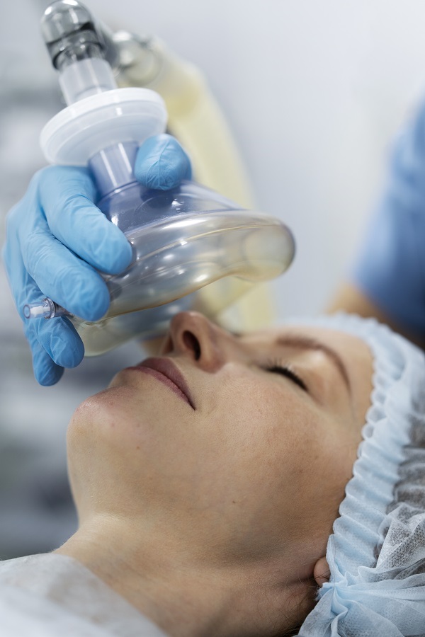 A patient lying on a surgical table with an anesthesia mask held by a medical professional wearing blue gloves.