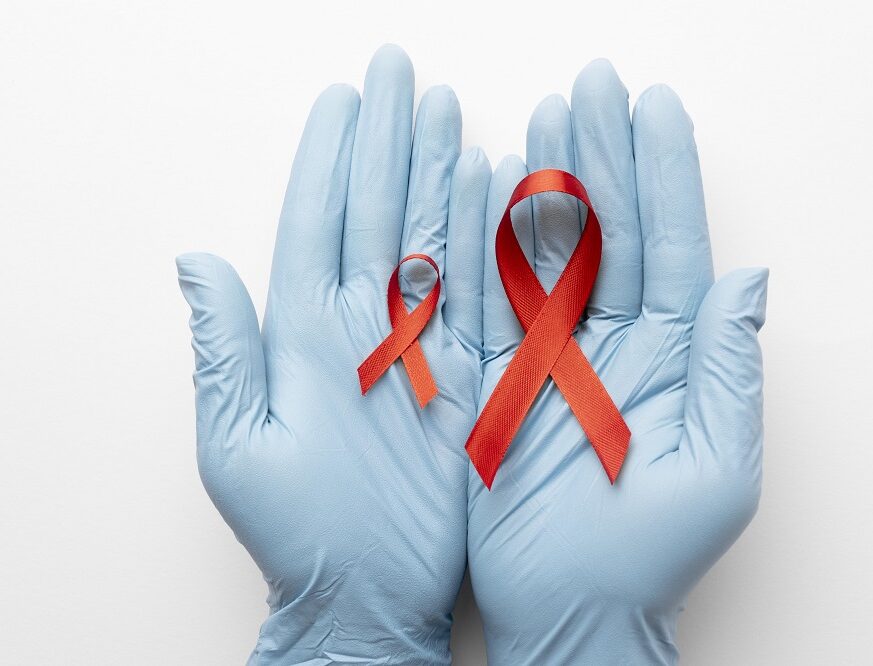 Two red AIDS awareness ribbons placed on blue-gloved hands symbolizing HIV/AIDS awareness and support.
