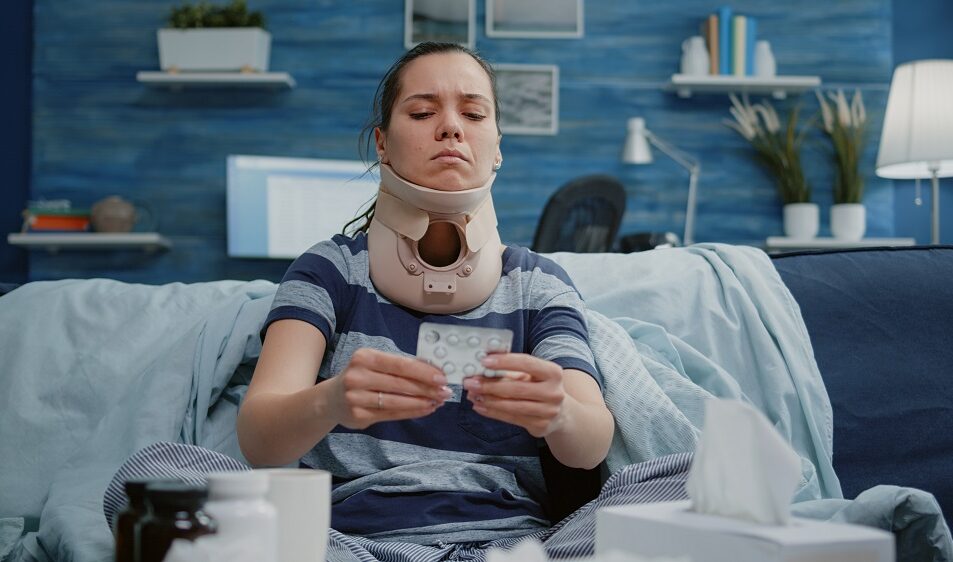 Injured woman with a cervical neck collar reading a medication label while sitting on a bed.