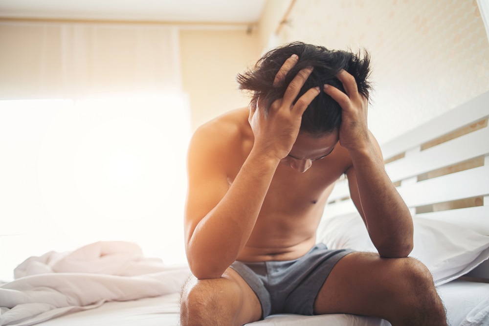 A shirtless man sitting on a bed with his head in his hands, appearing distressed or deep in thought, in a brightly lit bedroom.